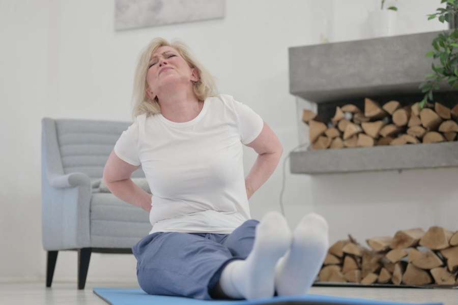 Femme assise sur un tapis de sol ressentant une vive douleur, illustrant le lien entre Yoga et mal de dos.