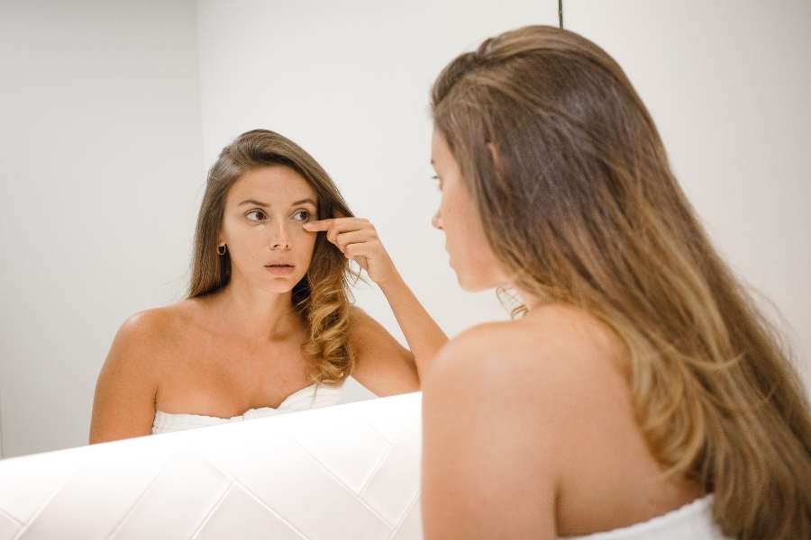Une jeune femme se regarde dans le miroir de la salle de bain et applique délicatement son anti-cernes sous l'œil avec son doigt.